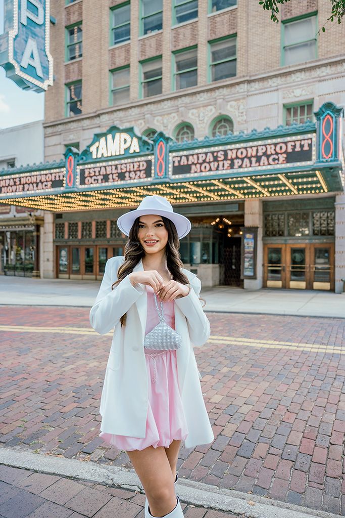 Stylish woman in pink dress, white blazer and hat poses before Tampa's Nonprofit Movie Palace theatre.