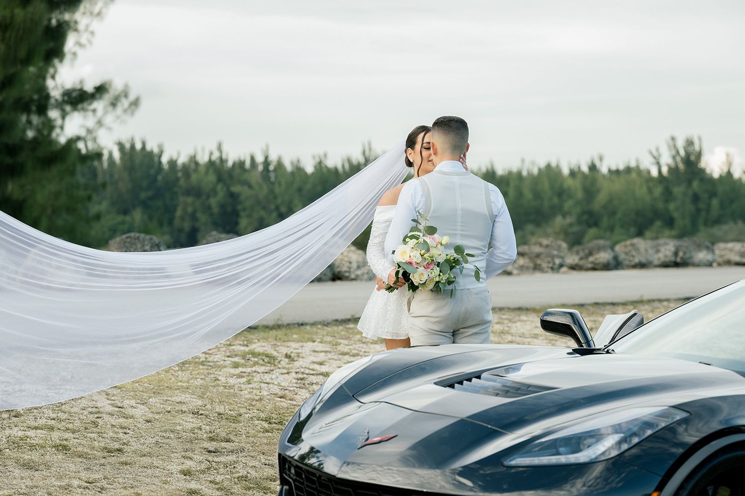 Bride and groom embrace beside a black sports car, her veil flowing in the wind, with lush trees in the background.