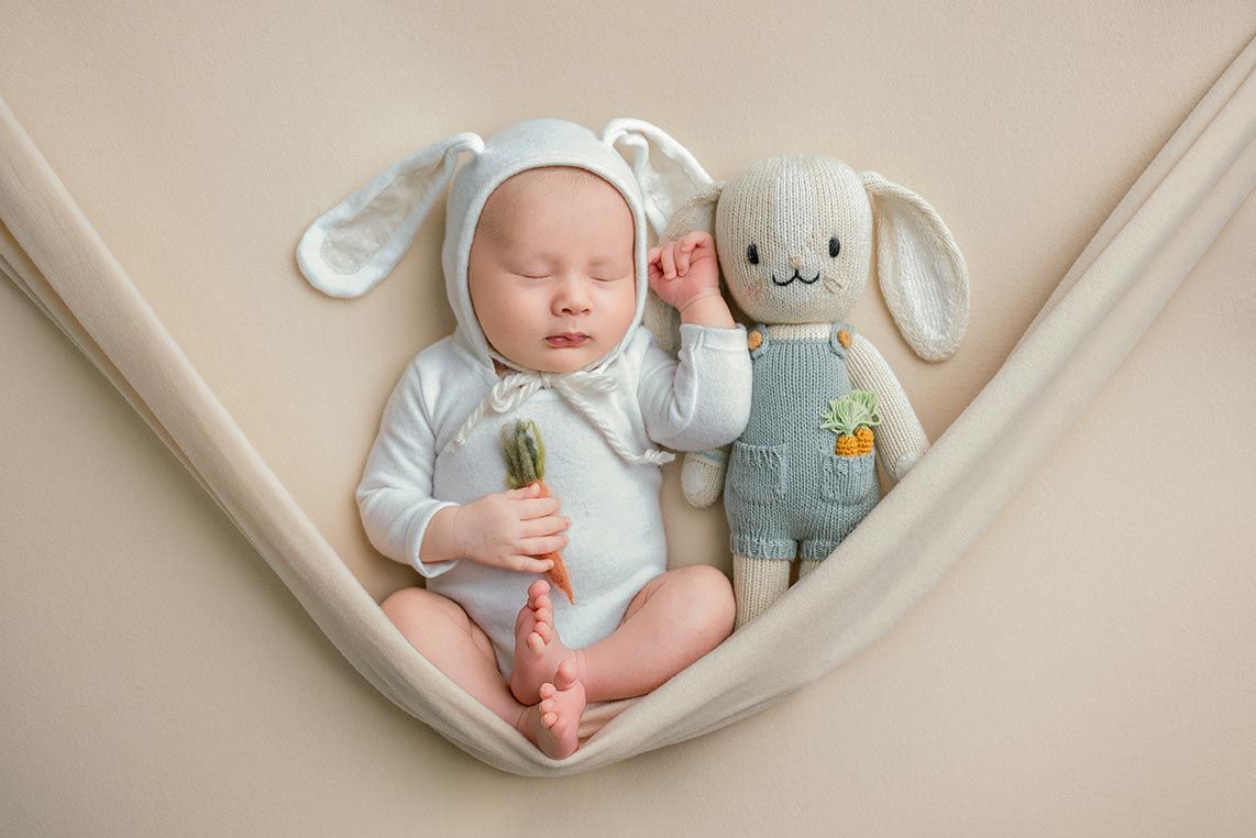 A sleeping baby in blue onesie lies in a hammock next to a plush bunny wearing denim overalls.