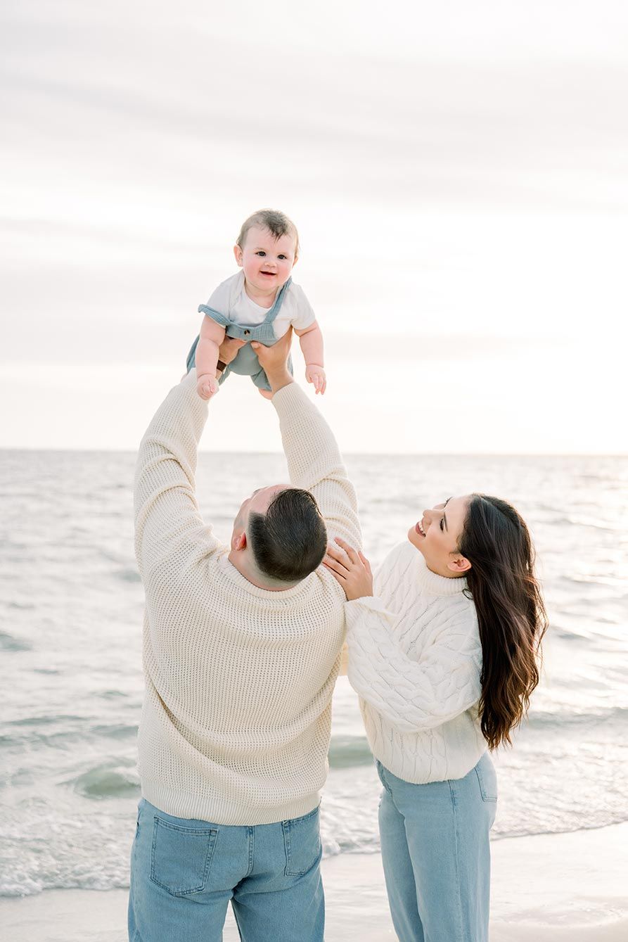 Family wearing coordinated white sweaters and jeans playing with baby at sunny beach.