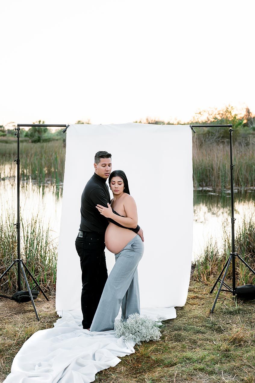 A bride and groom share an intimate moment behind a white backdrop in an outdoor setting with tall grass and natural light.