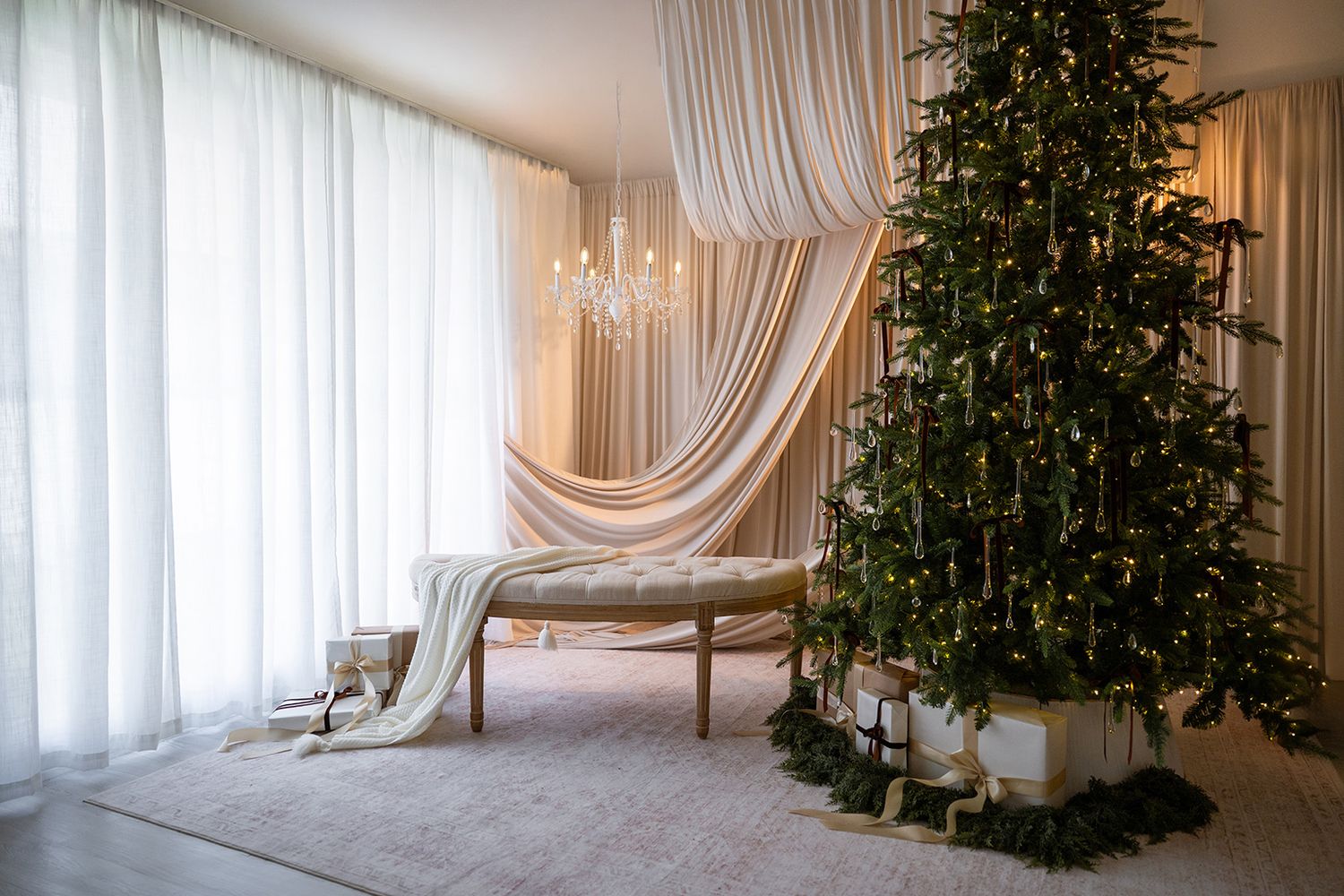 Elegant Christmas tree with lights and white gifts beside a chaise lounge, chandelier, and draped white curtains.