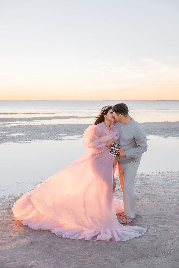 Couple in pink gown and gray suit share a tender moment on a beach at sunset, holding an ultrasound photo.