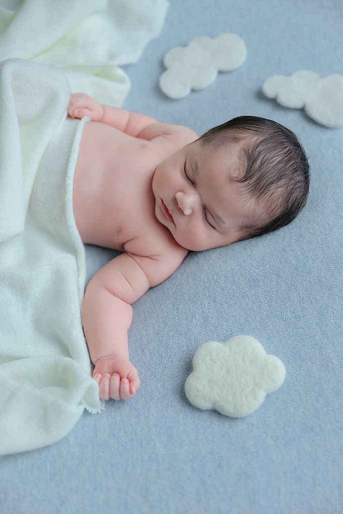 Smiling newborn baby sleeping on a gray blanket with white cloud decorations and a light green wrap.