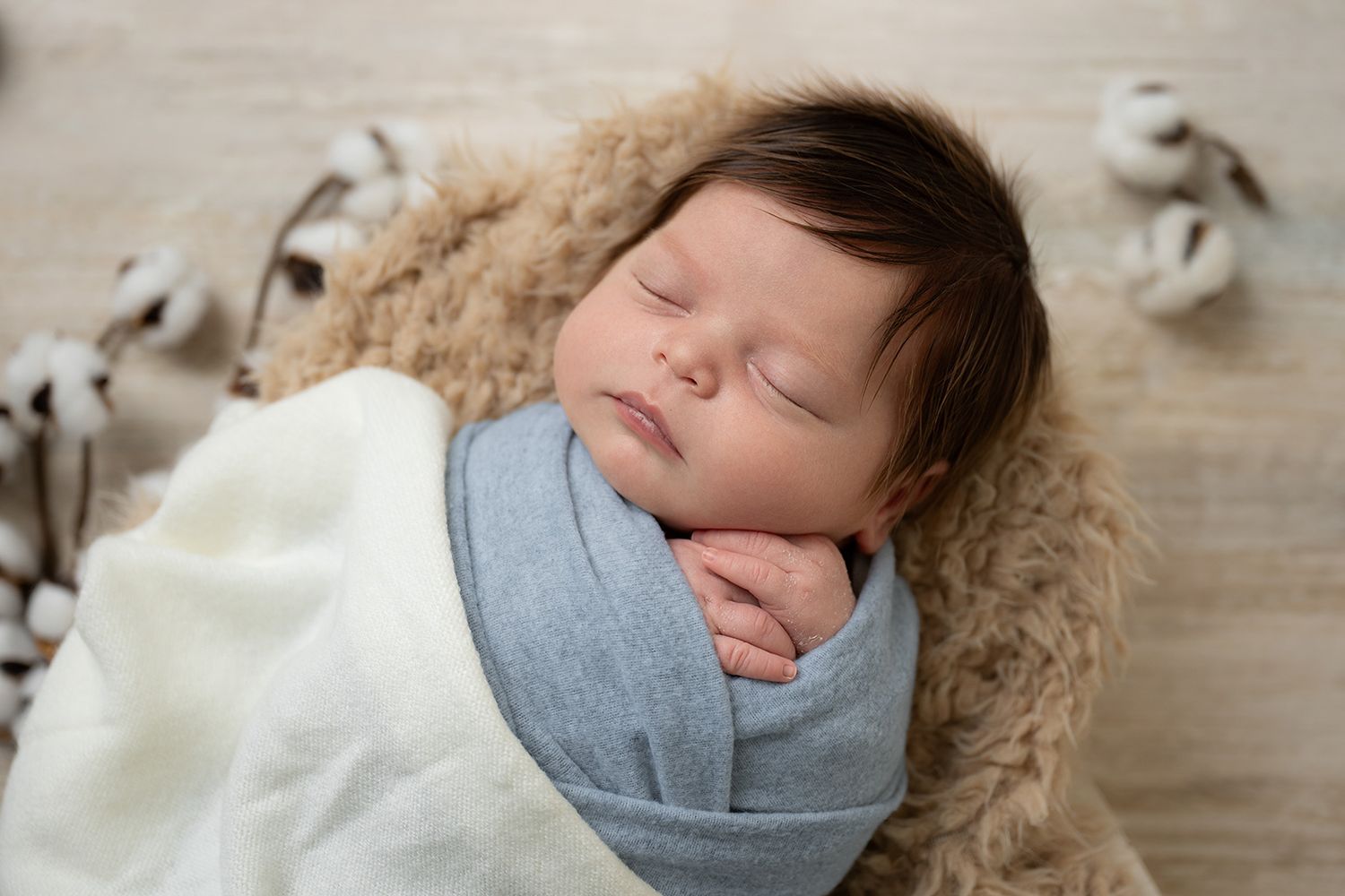 Peaceful newborn baby swaddled in a soft blue wrap, sleeping on a cozy tan fur blanket with cotton stems nearby.