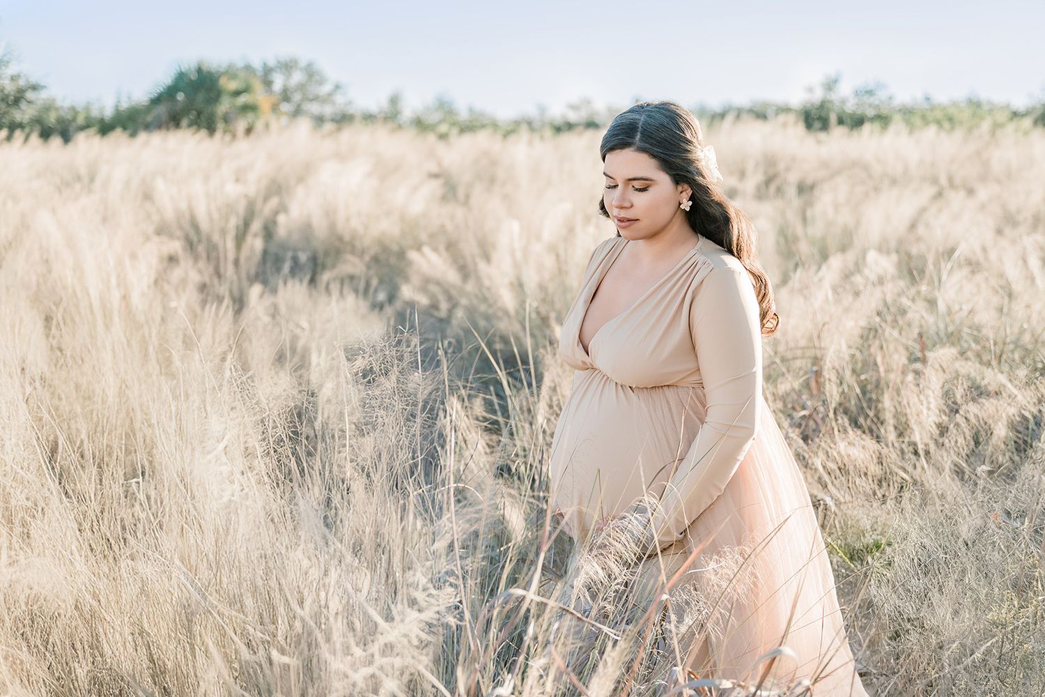 Pregnant woman in beige dress standing in golden grass field during golden hour maternity photo session.