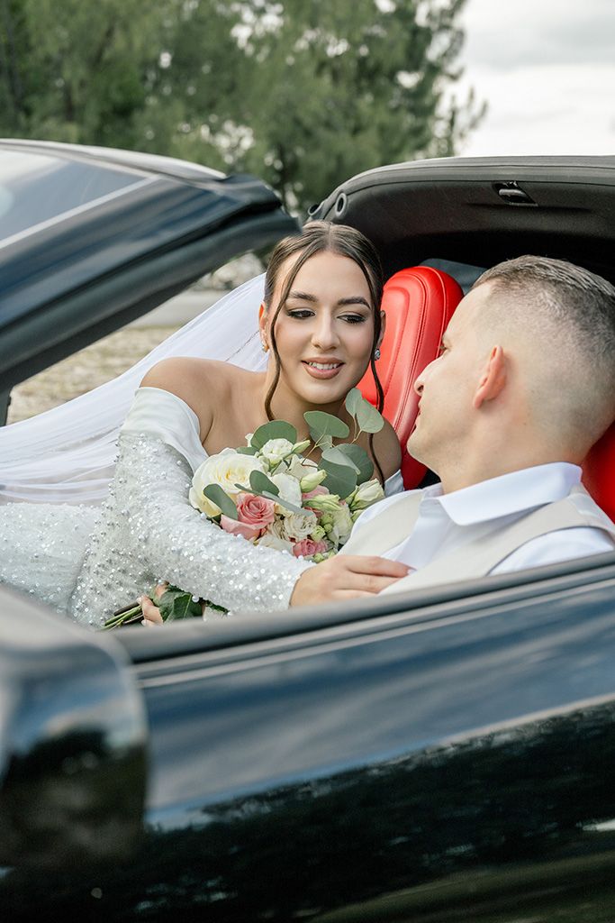 Bride and groom share a romantic moment in a black convertible car with red interior, bride holding a bouquet.