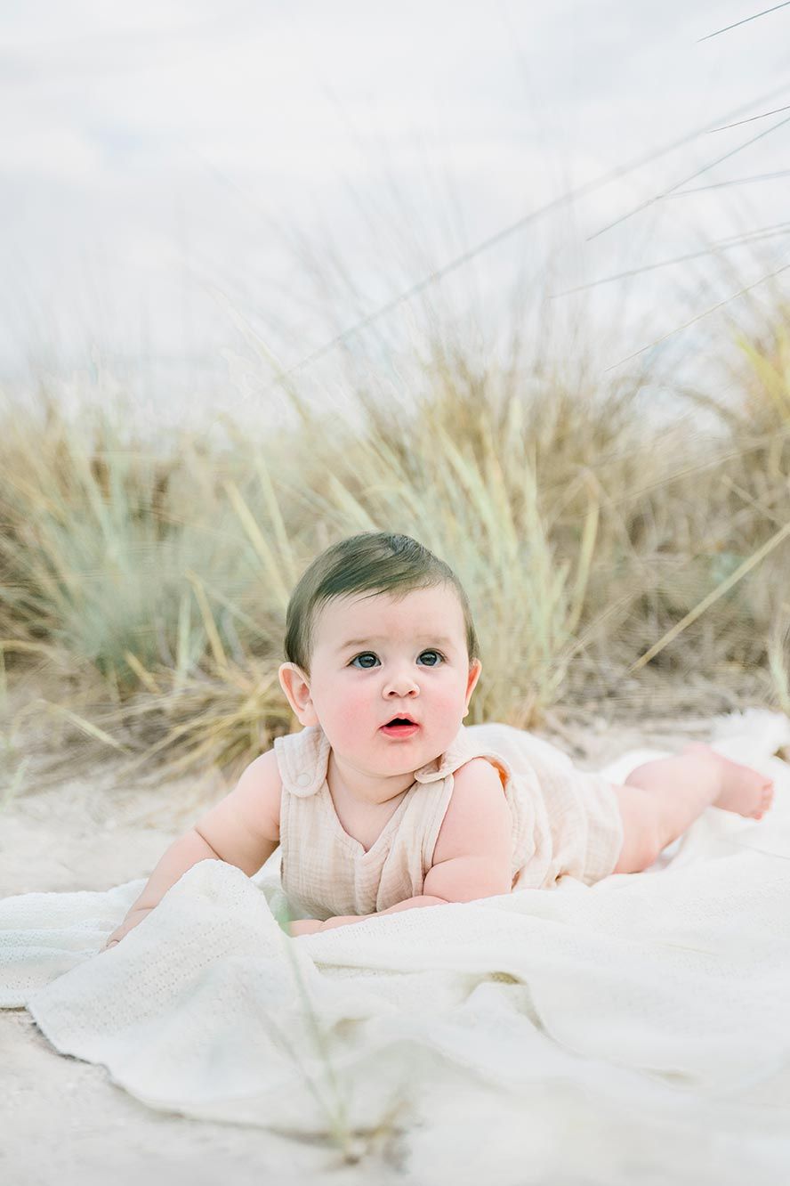 Baby lying on white blanket at beach with sea grass in soft dreamy lighting.