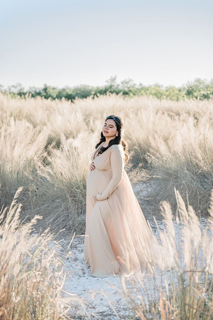 Elegant pregnant woman in blush pink flowing gown standing gracefully among tall golden grass in soft natural light.