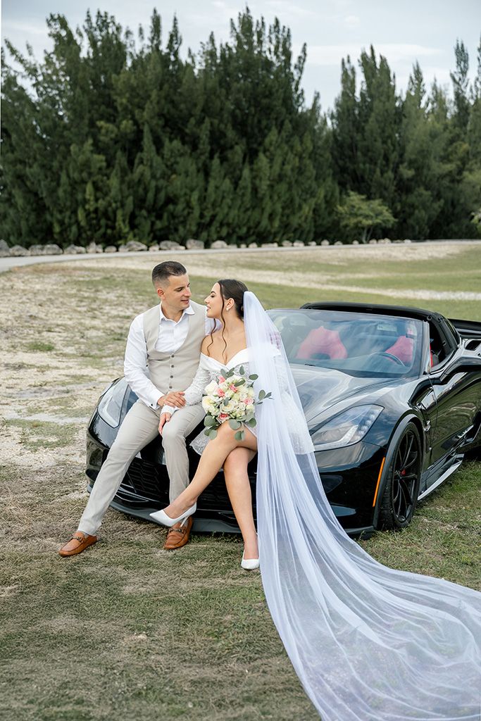 Bride and groom posing elegantly on a black sports car with a flowing veil in an outdoor setting.