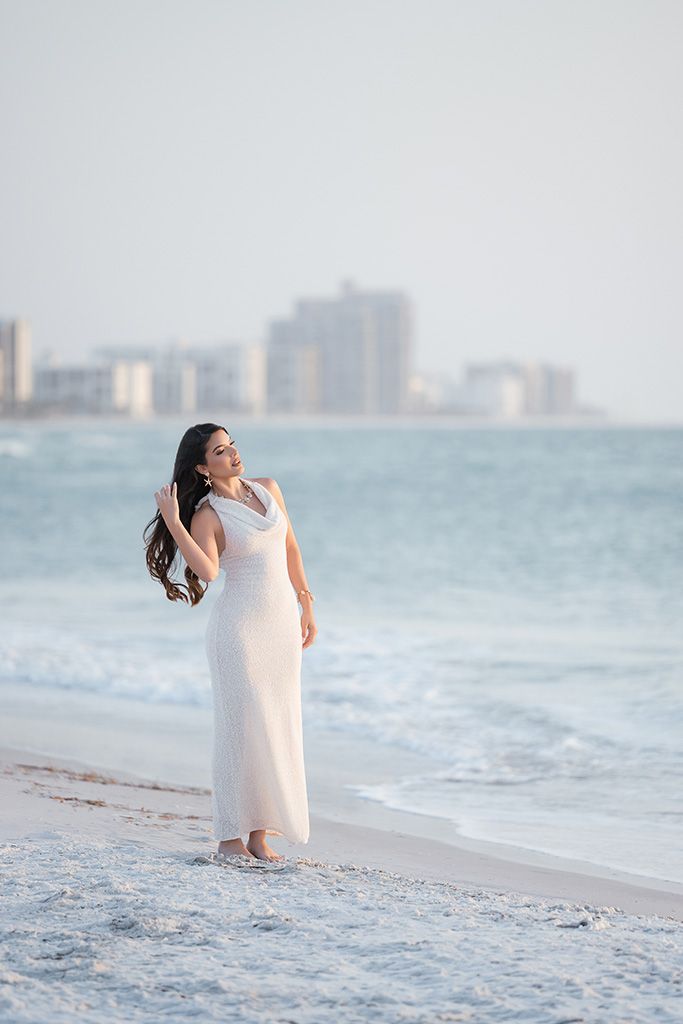 Woman in flowing white gown standing on shoreline with ocean waves and city skyline backdrop.