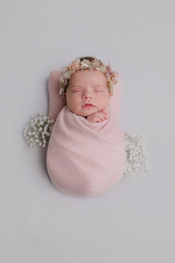 Sleeping newborn baby swaddled in pink wrap with floral crown and baby's breath flowers on white background.