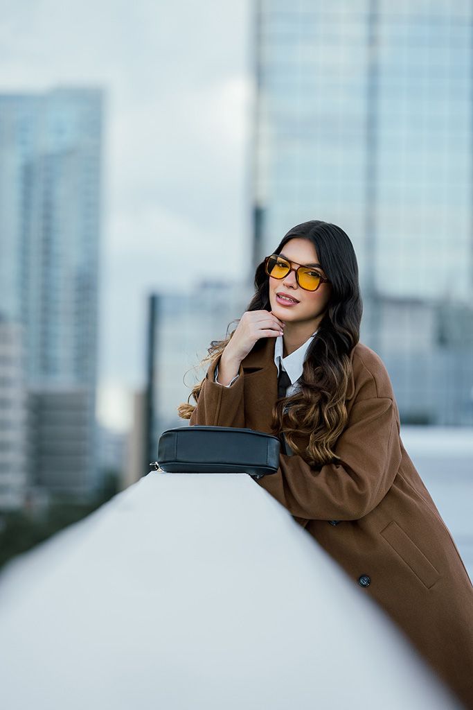 Fashion woman in brown coat and yellow sunglasses leans on white rooftop ledge with city skyline background.
