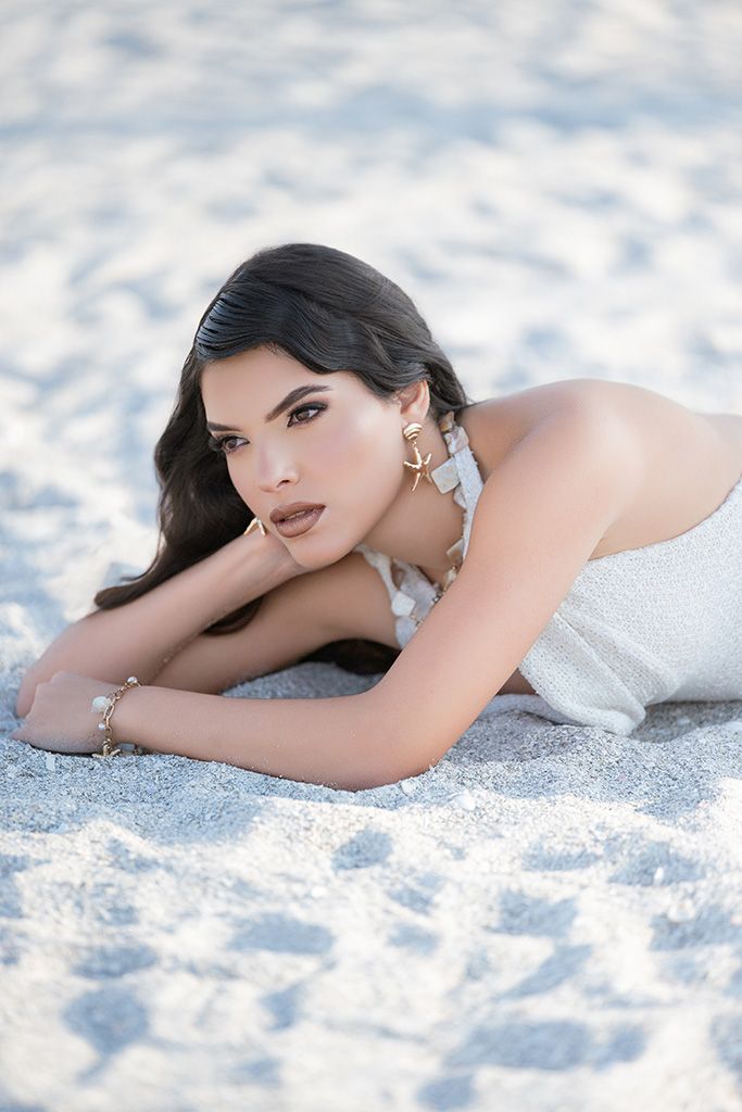 A woman with dark hair lies on white sand, wearing a silver dress and gold earrings, gazing at the camera.