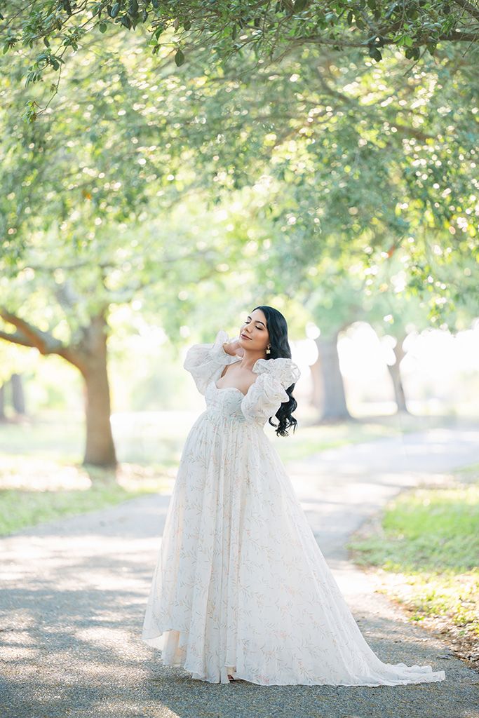 A bride in a flowing white lace gown with puff sleeves poses gracefully on a tree-lined path in soft natural light.