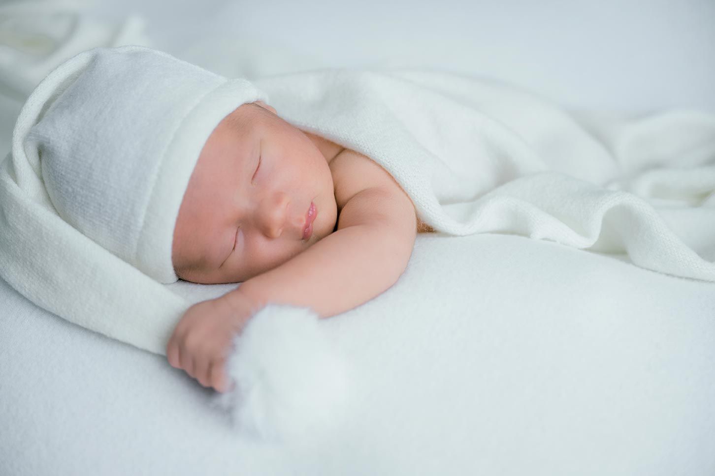 Peaceful newborn baby sleeping wrapped in a soft white blanket wearing a white knit hat on a white background.