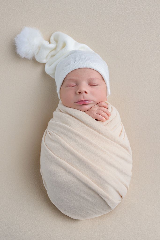 Peaceful newborn baby swaddled in cream wrap wearing white pom-pom hat, sleeping soundly on beige background.