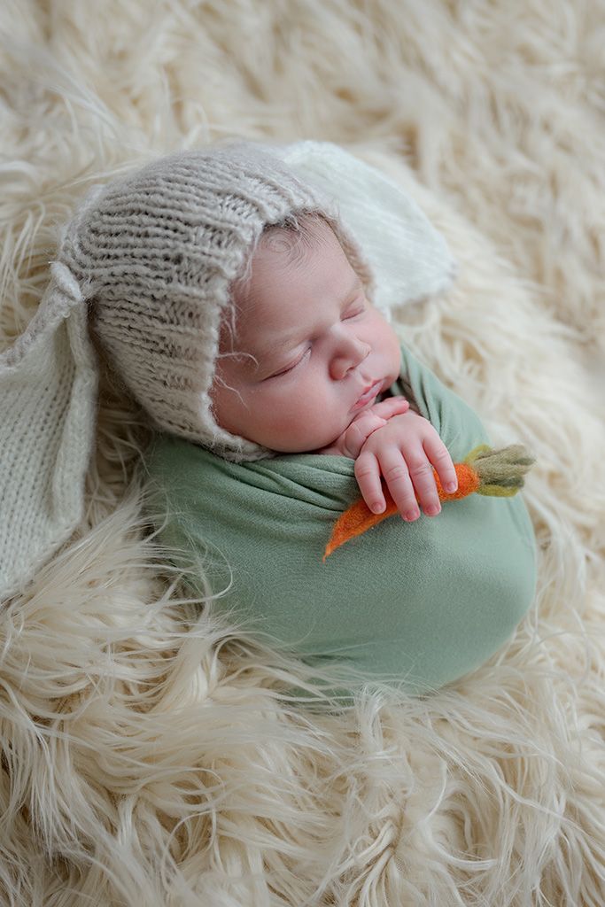 Sleeping newborn baby wrapped in green swaddle, wearing knit bunny ear bonnet, holding a tiny carrot, on fluffy cream rug.
