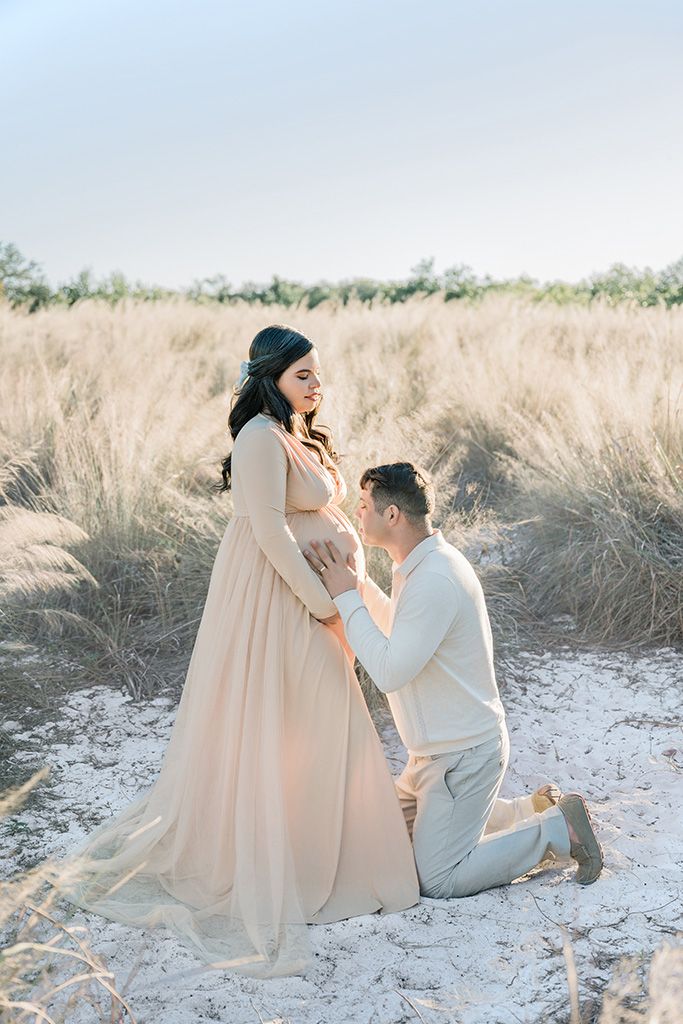Pregnant woman in blush gown with man kneeling before her in a golden field during a maternity photoshoot.