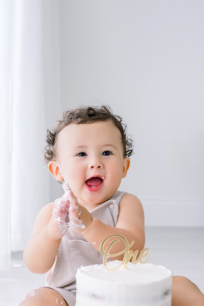 A joyful baby celebrates their first birthday smashing a white cake with a gold “One” topper, hands covered in frosting.