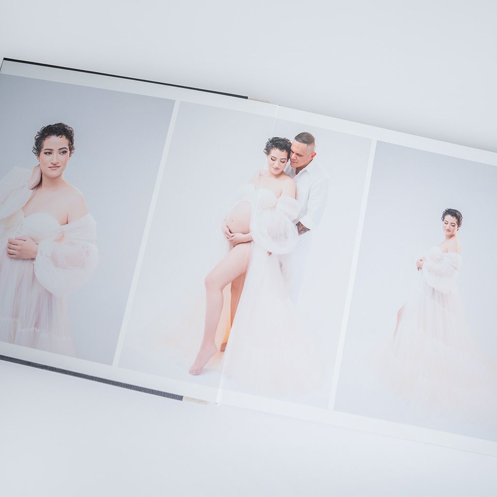 Open photo book displaying elegant maternity portraits of a pregnant woman in a white dress with her partner.
