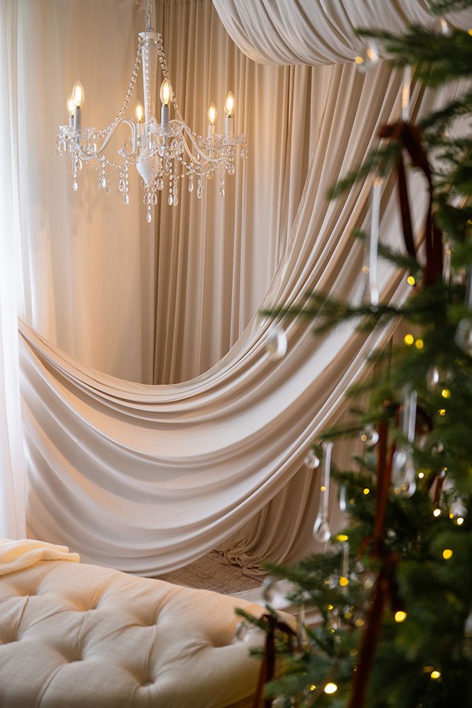 Elegant white draped curtains with crystal chandelier and Christmas tree with warm lights in luxury interior.