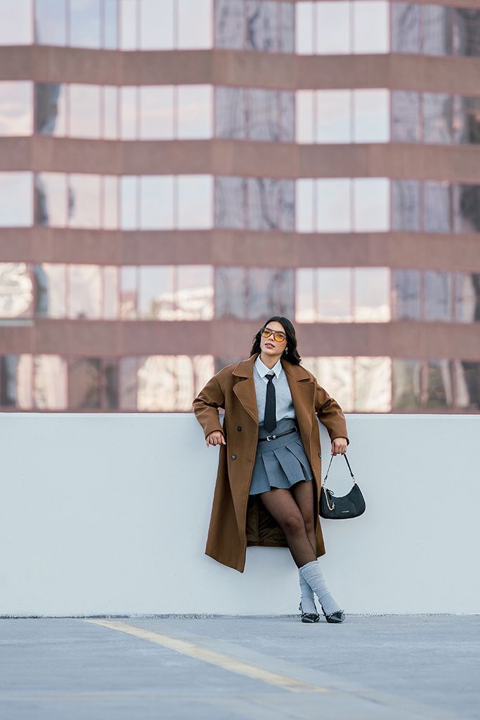 Stylish woman in brown coat, gray pleated skirt, and tie leans against white wall holding black bag outdoors.