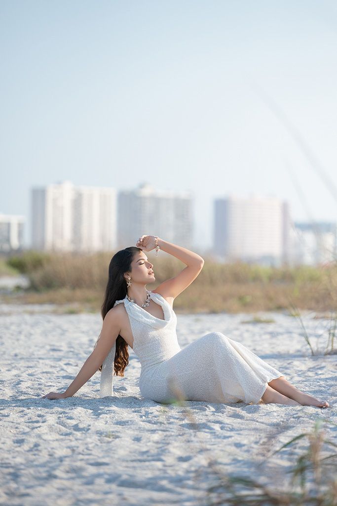 Woman in white dress sitting gracefully on sandy beach with coastal buildings in background.