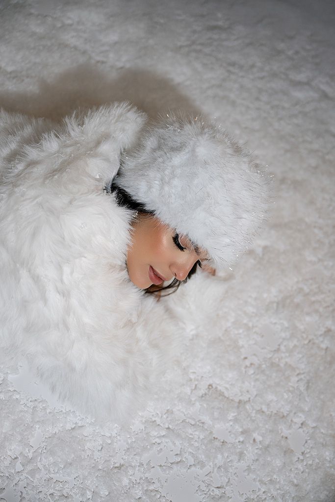 Woman lying in snow wearing a white faux fur coat and hat, peeking out from the fluffy white material.