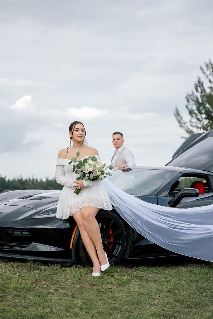 Bride in white dress holds bouquet beside black Corvette, groom in white shirt stands behind car outdoors.
