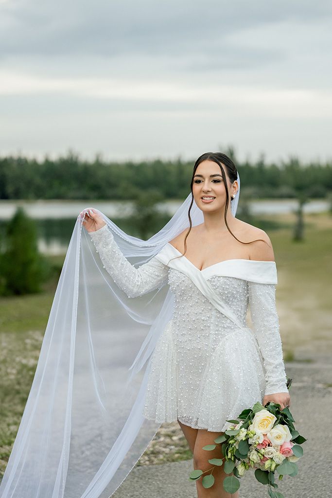 Smiling bride in short white lace off-shoulder gown holds bouquet and veil outdoors near lake.