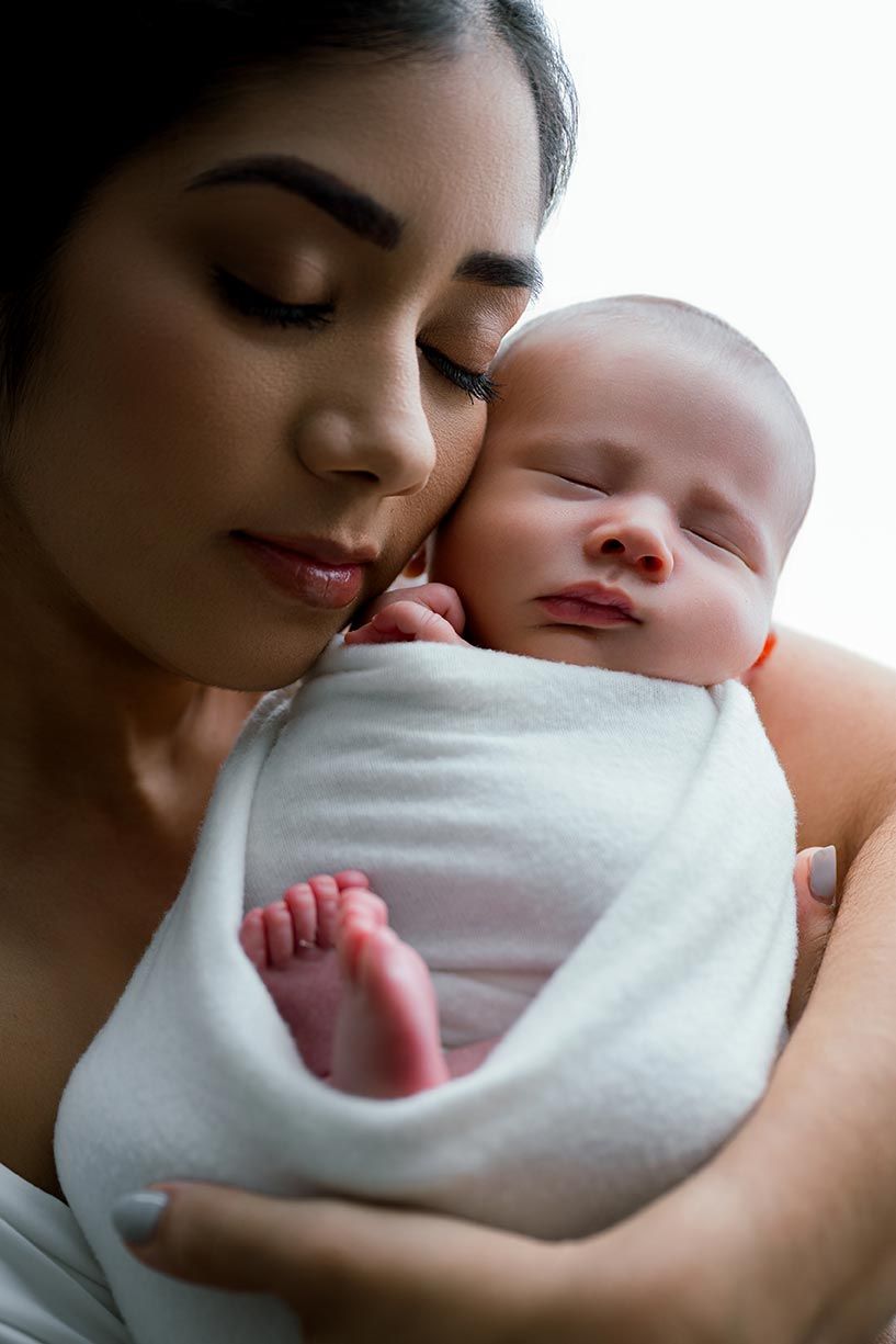 Mother gently kissing sleeping swaddled newborn baby wrapped in white cloth with tiny feet visible.