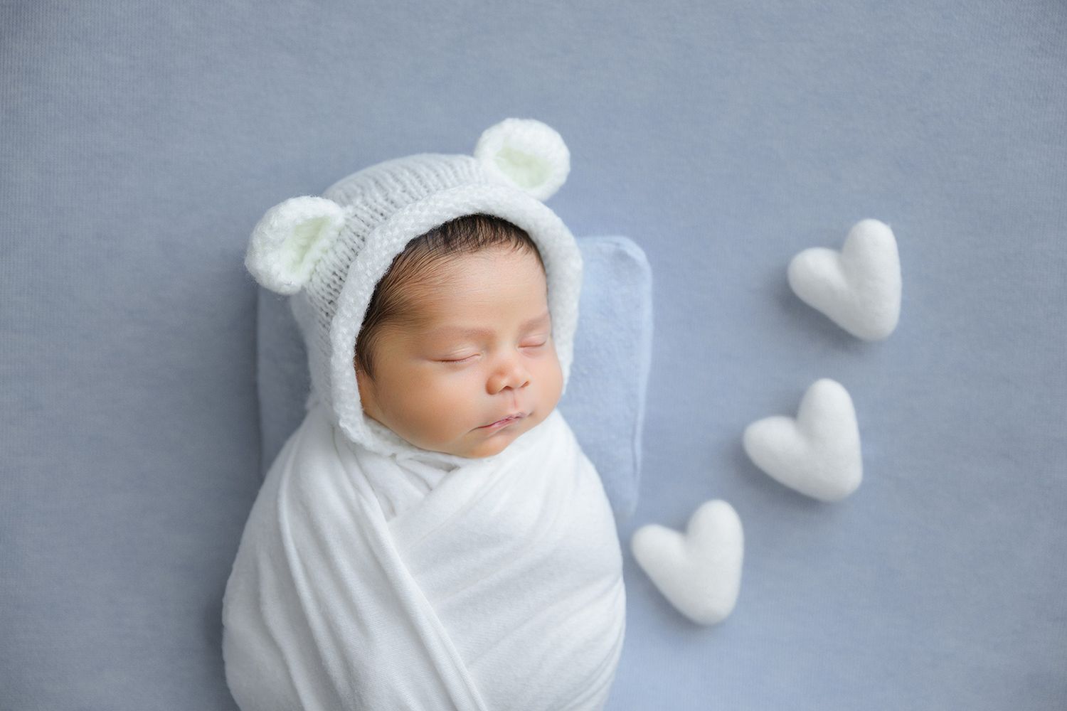 Sleeping newborn baby swaddled in white wrap wearing a knit bear bonnet on a blue background with heart props.