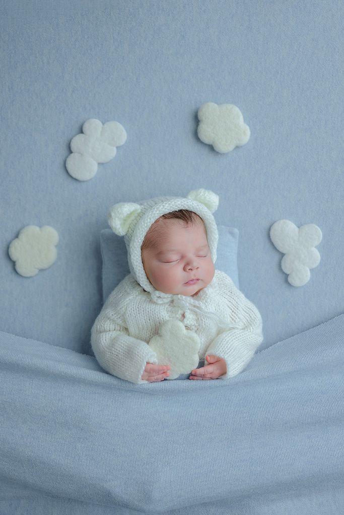 Sleeping newborn baby in white knit bear outfit on blue blanket with felt cloud decorations.