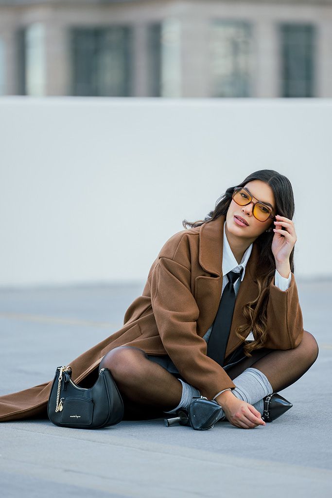 Woman in brown coat, gray skirt, tie, and orange glasses sits cross-legged on rooftop holding black handbag.