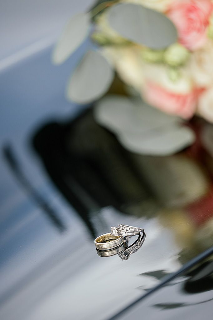 Two wedding rings stacked on a reflective surface with a blurred floral bouquet in the background.