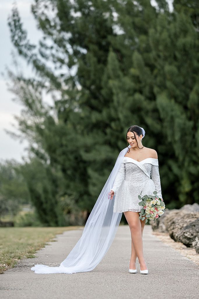 Bride in short white beaded off-shoulder gown with flowing veil holds bouquet on outdoor path with tall green trees.