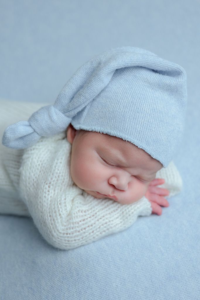 Sleeping newborn baby wearing a white knit outfit and light blue knotted hat on a soft blue blanket.