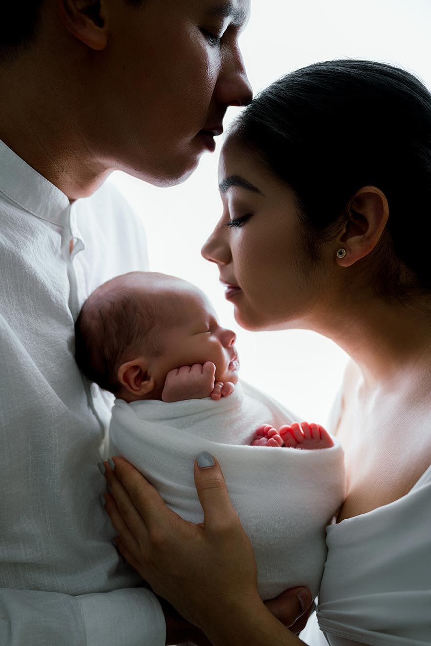 Parents tenderly kissing their newborn baby wrapped in white swaddle in a softly lit intimate portrait.