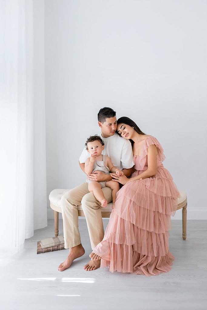 A family of three poses in a bright studio, mother in pink tulle gown, father kissing toddler son on a bench.