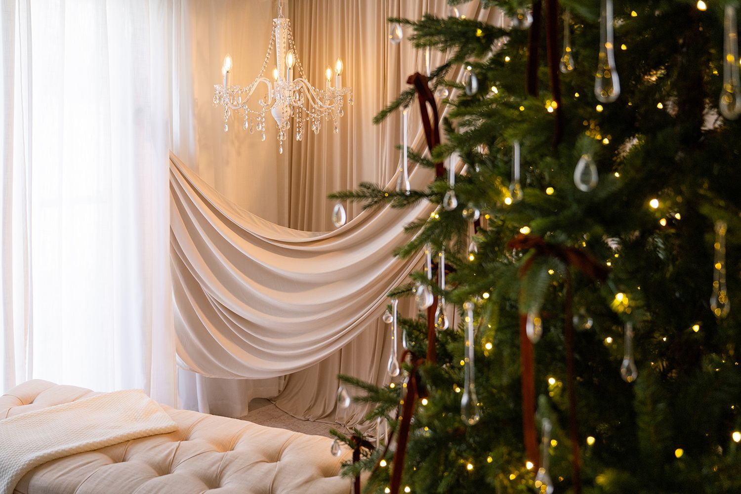 Festive room featuring a decorated Christmas tree, crystal chandelier, cream curtains, and a tufted white bench.