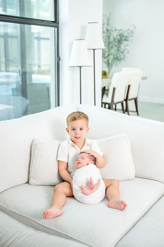 A young toddler boy sits on a white sofa gently holding a swaddled newborn baby sibling in a bright modern living room.