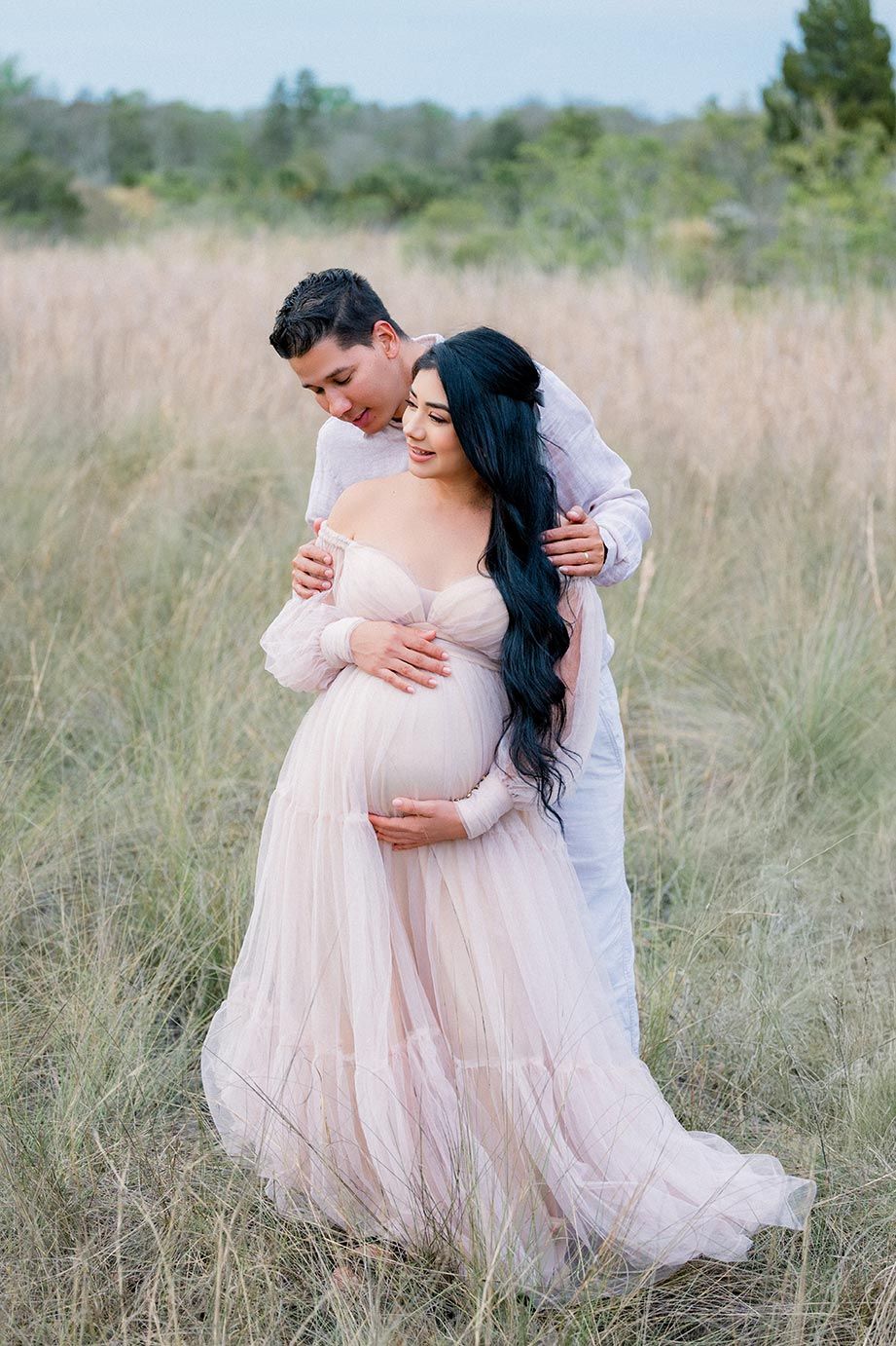 A romantic maternity photo in a field shows a couple embracing, with the woman wearing a flowing blush pink gown.