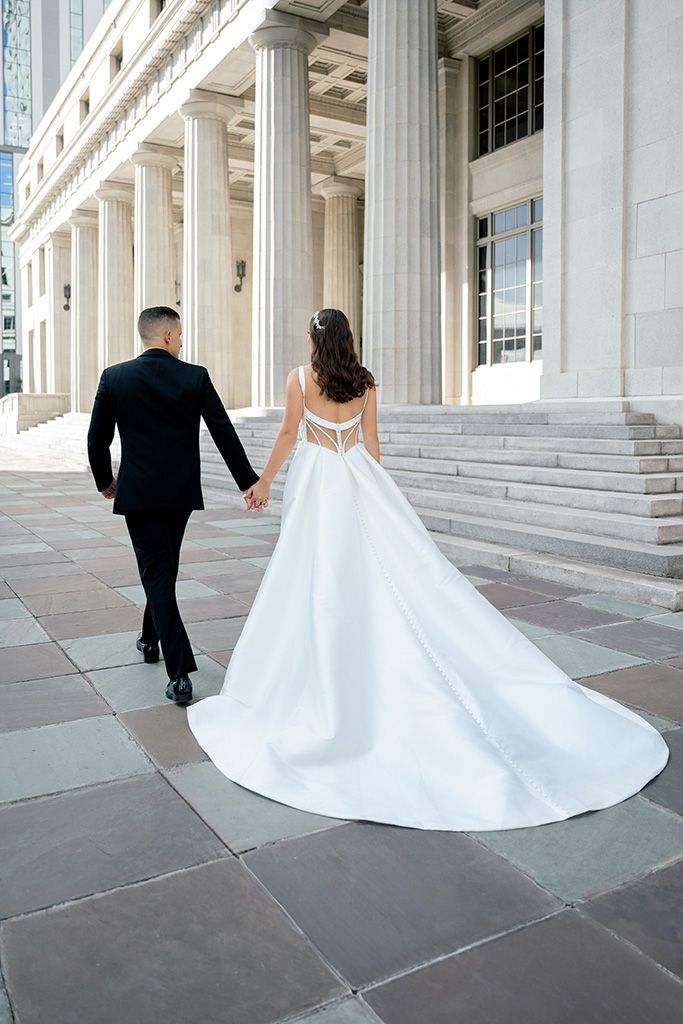 Bride in elegant white gown with train holds groom's hand walking toward grand columned building.