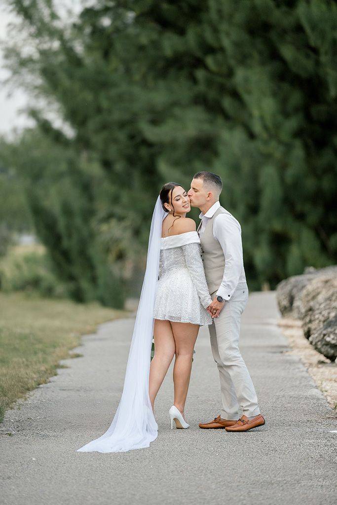 Bride and groom share a kiss on a tree-lined path, bride wearing short white dress with long veil.