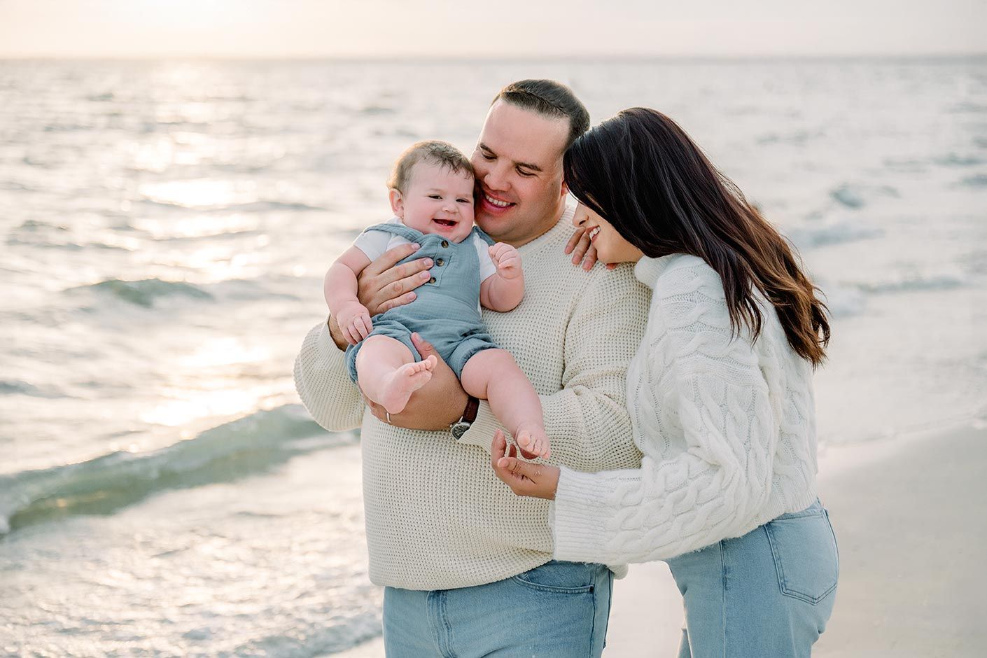 Loving parents cuddling laughing baby on sandy beach with gentle ocean waves and sunset in background.