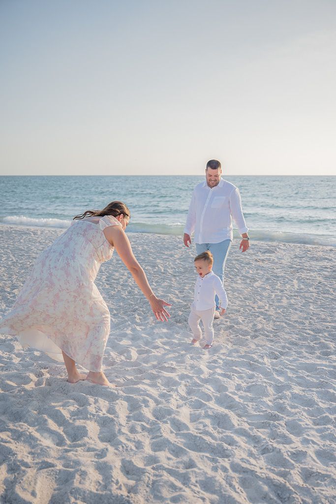 Family of three playing on white sand beach at sunset, mother reaching for toddler while father watches.