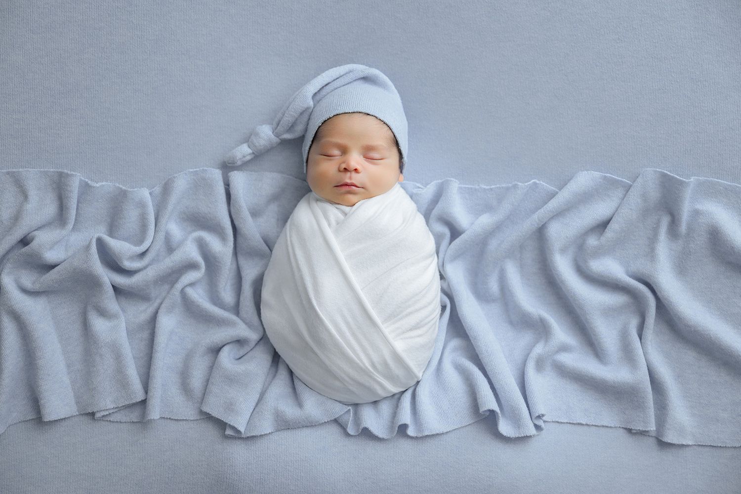Peaceful newborn baby swaddled in white wrap wearing a blue knot hat on a soft gray blanket.