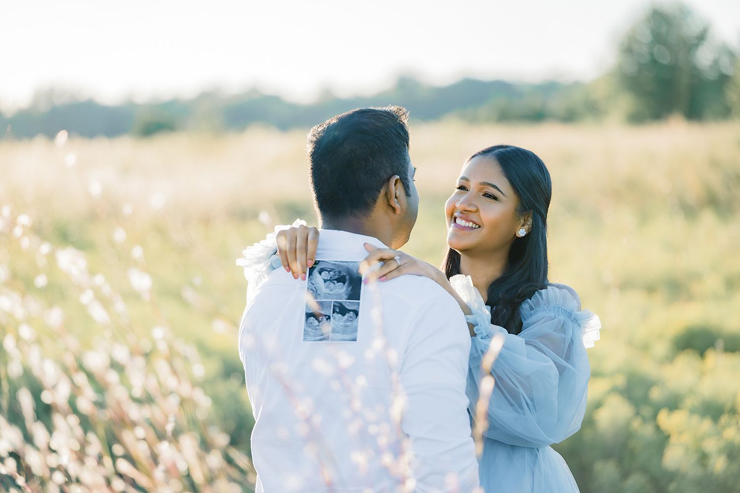 A smiling couple embraces in a sunlit flower field, the woman in blue gazing lovingly at her partner.