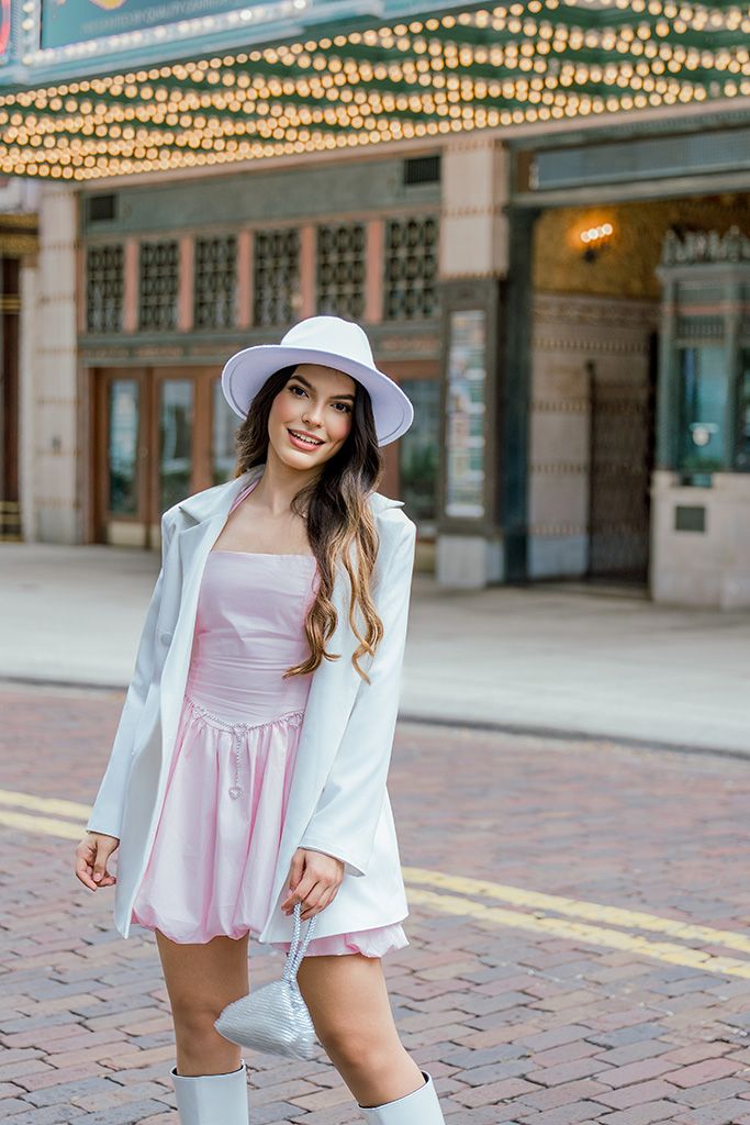 Woman in pink mini dress, white blazer, hat, and boots posing outside a theater with marquee lights.